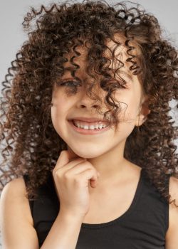 Close up portrait of adorable smiling girl in black sports top posing, isolated on gray studio background. Little female with curly brunette hair on face looking at camera and touching neck.