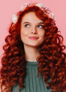 Young girl with red hair and flowers on her head crosses fingers. Portrait of blue-eyed woman in green dress on pink background